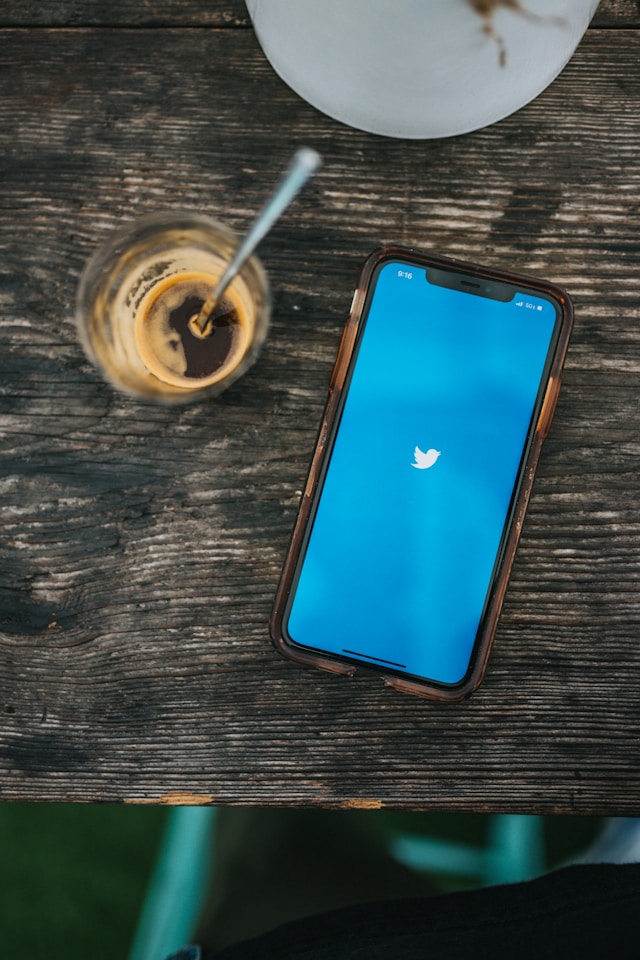 Um smartphone exibindo o logotipo do Twitter ao lado de uma xícara de café em uma mesa de madeira.