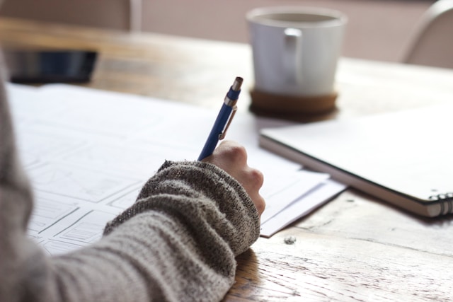 Une personne portant un pull beige écrit dans un livre à côté d'une tasse en céramique blanche.